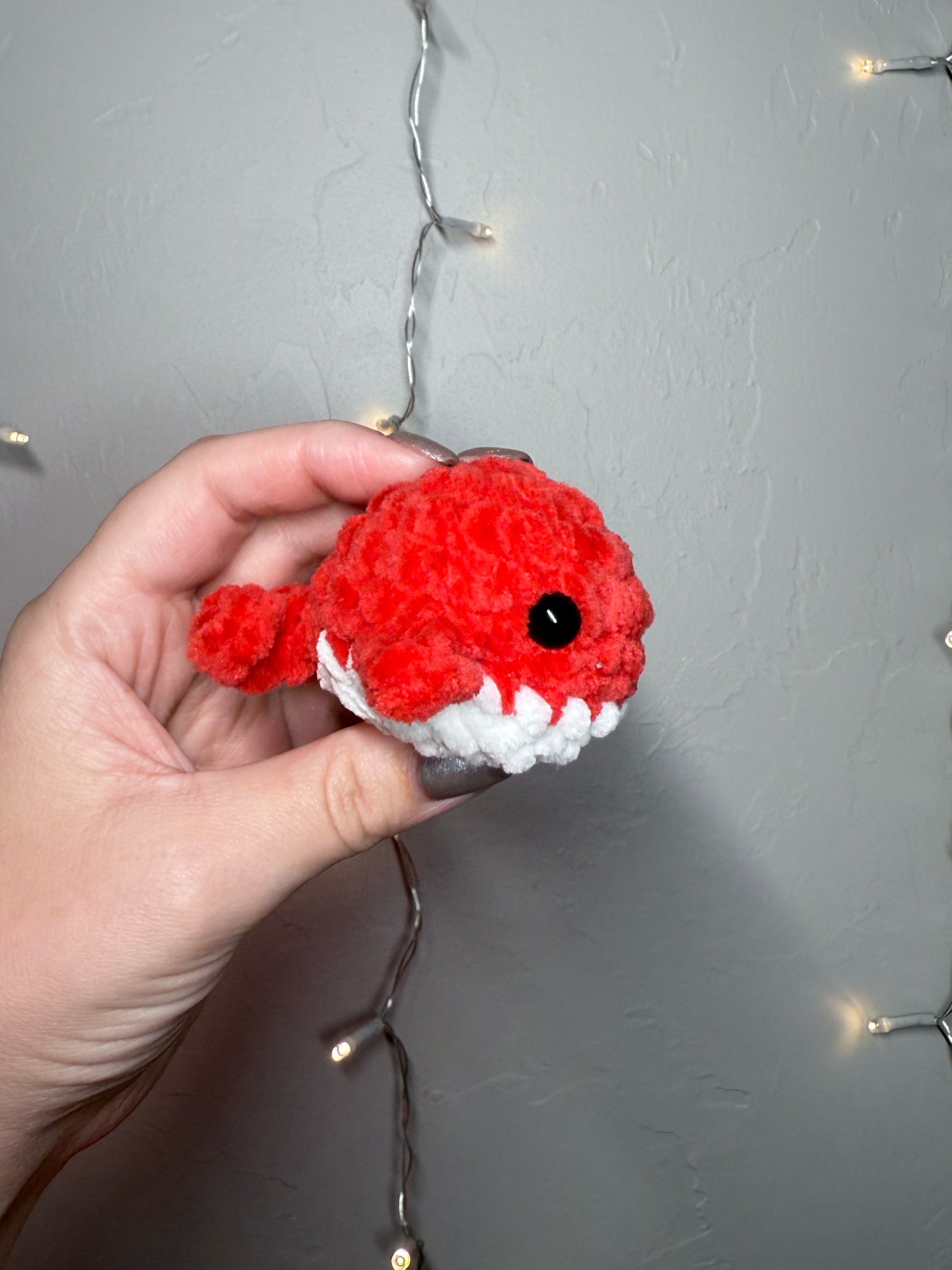 Hand holding a small red and white crocheted fish toy against a gray background with string lights.