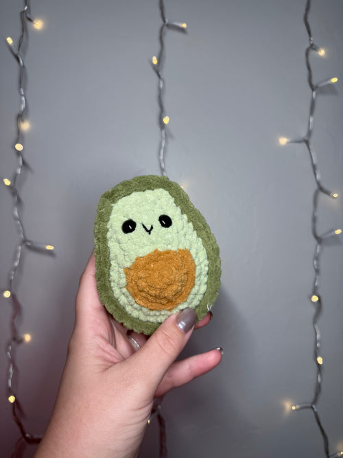 Hand holding a knitted avocado toy with a neutral background and string lights.